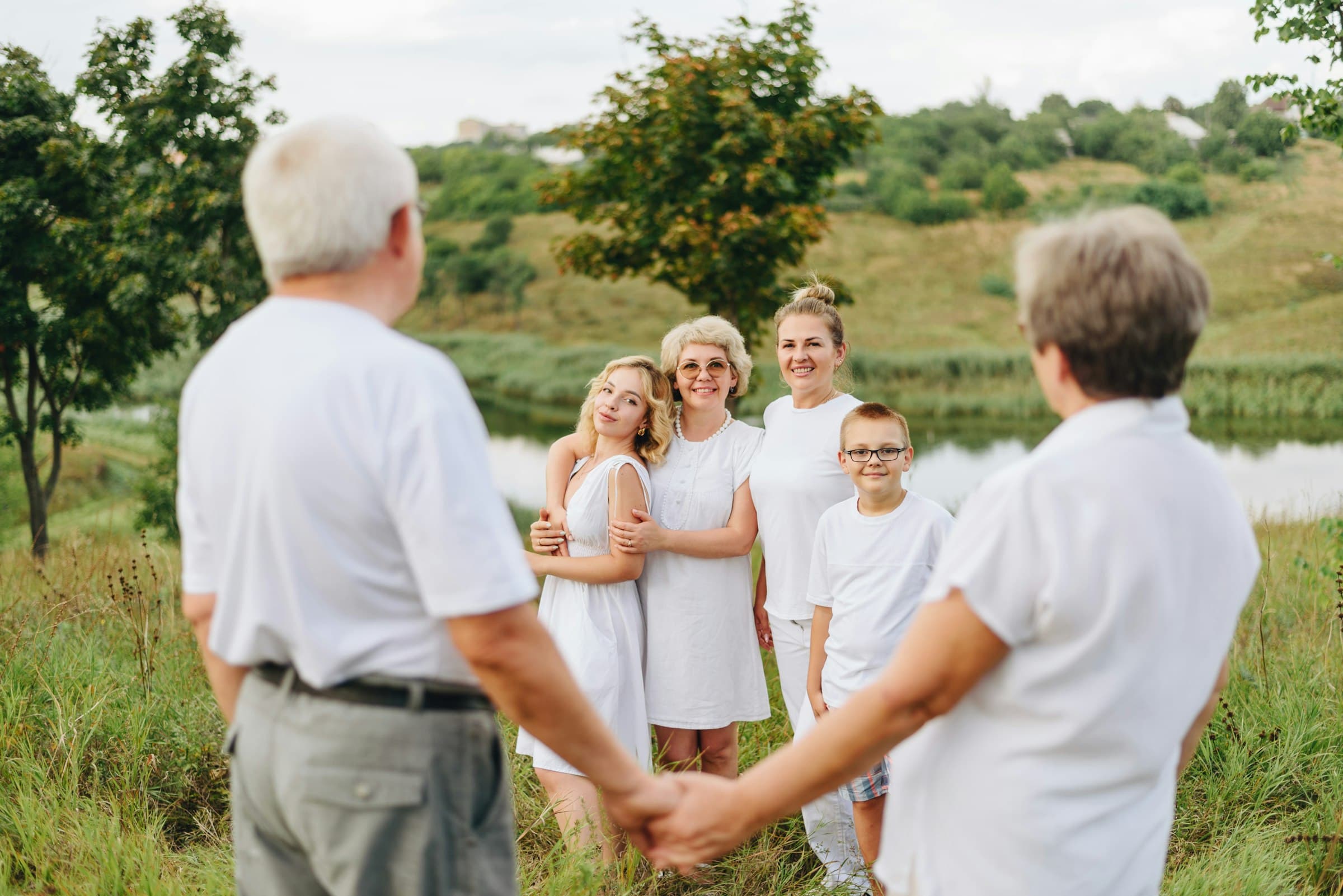 An older couple seen from behind holding hands, facing an older woman, two younger women and a young boy gathered on a grassy riverbank